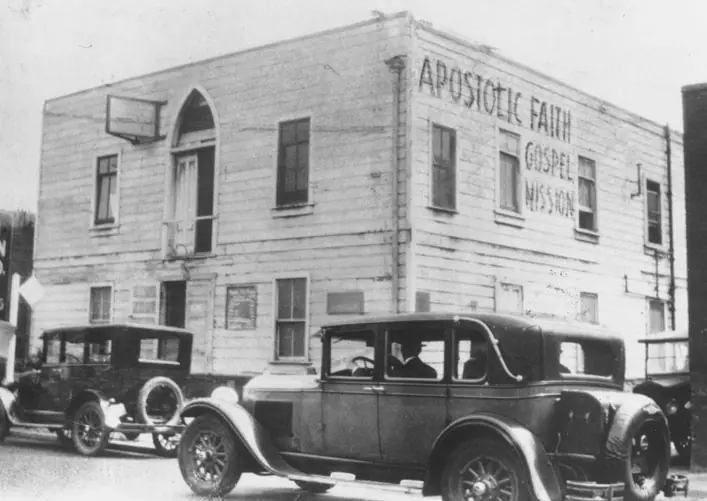 A two-storey white clapboard building with an arched upper window and the painted words Apostolic Faith Gospel Mission, with 1920s cars parked outside