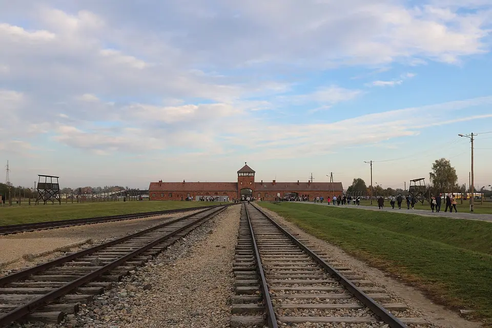 Railway tracks leading to the long brick gatehouse and central tower of Auschwitz II–Birkenau, with wooden watchtowers and visitors walking beside the line