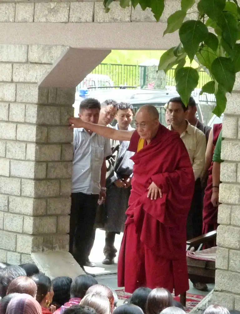 The Dalai Lama in maroon robes stepping through a stone doorway, one hand raised, with attendants behind him and a seated crowd in the foreground