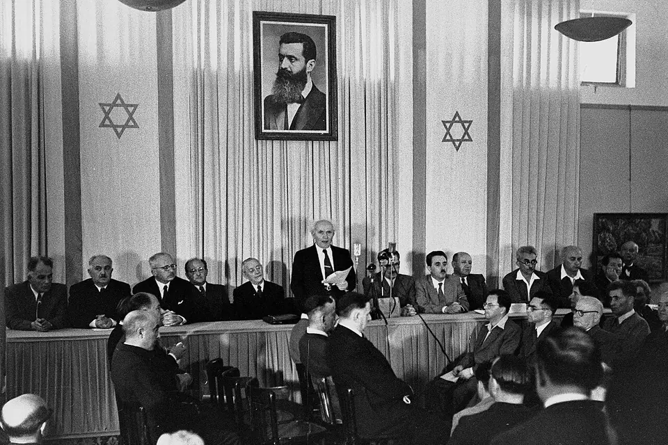 David Ben-Gurion standing at a long table of seated men, reading the declaration of the State of Israel beneath a large portrait of Theodor Herzl and two hanging flags with the Star of David
