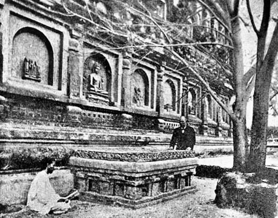 Old photograph of a man in dark clothes standing behind the carved stone Vajrāsana platform beside the Mahabodhi temple wall at Bodh Gaya