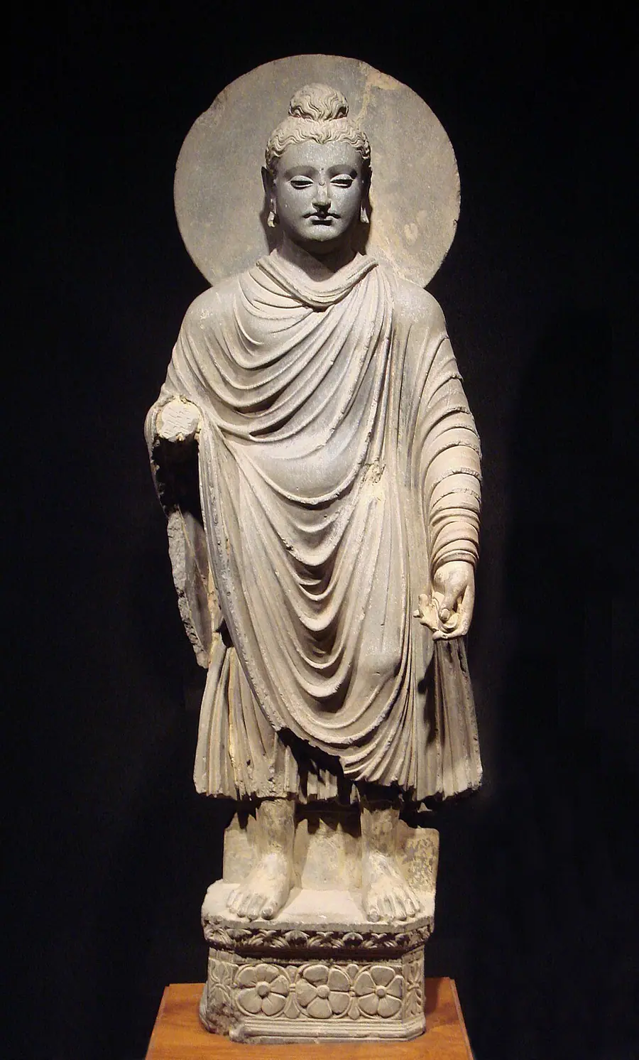 Grey schist standing Buddha from Gandhāra with a halo, topknot, moustache and heavy pleated robe in Hellenistic style