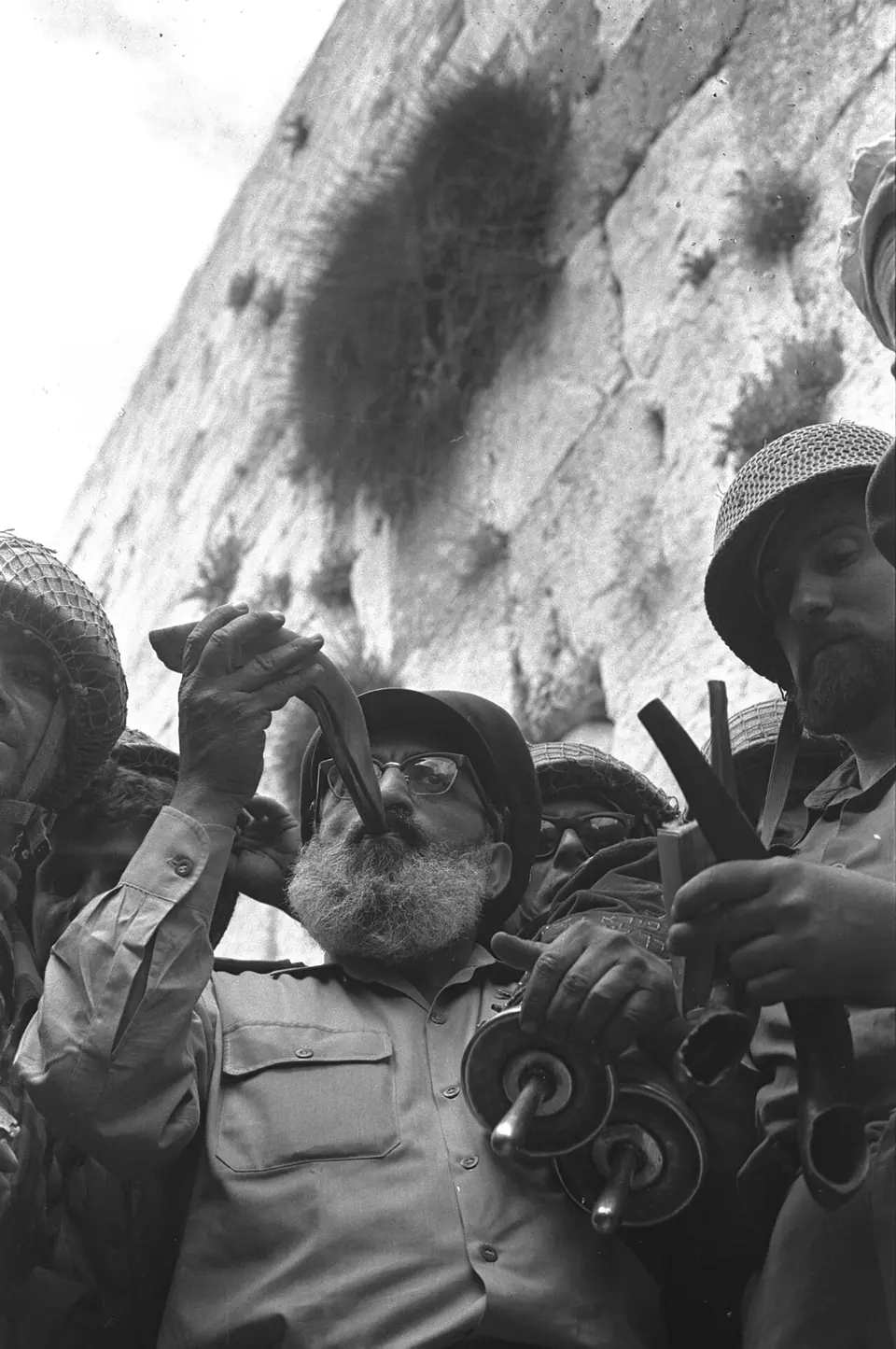 Rabbi Shlomo Goren in army uniform blowing a shofar and holding a Torah scroll, surrounded by helmeted Israeli soldiers at the foot of the Western Wall, June 1967