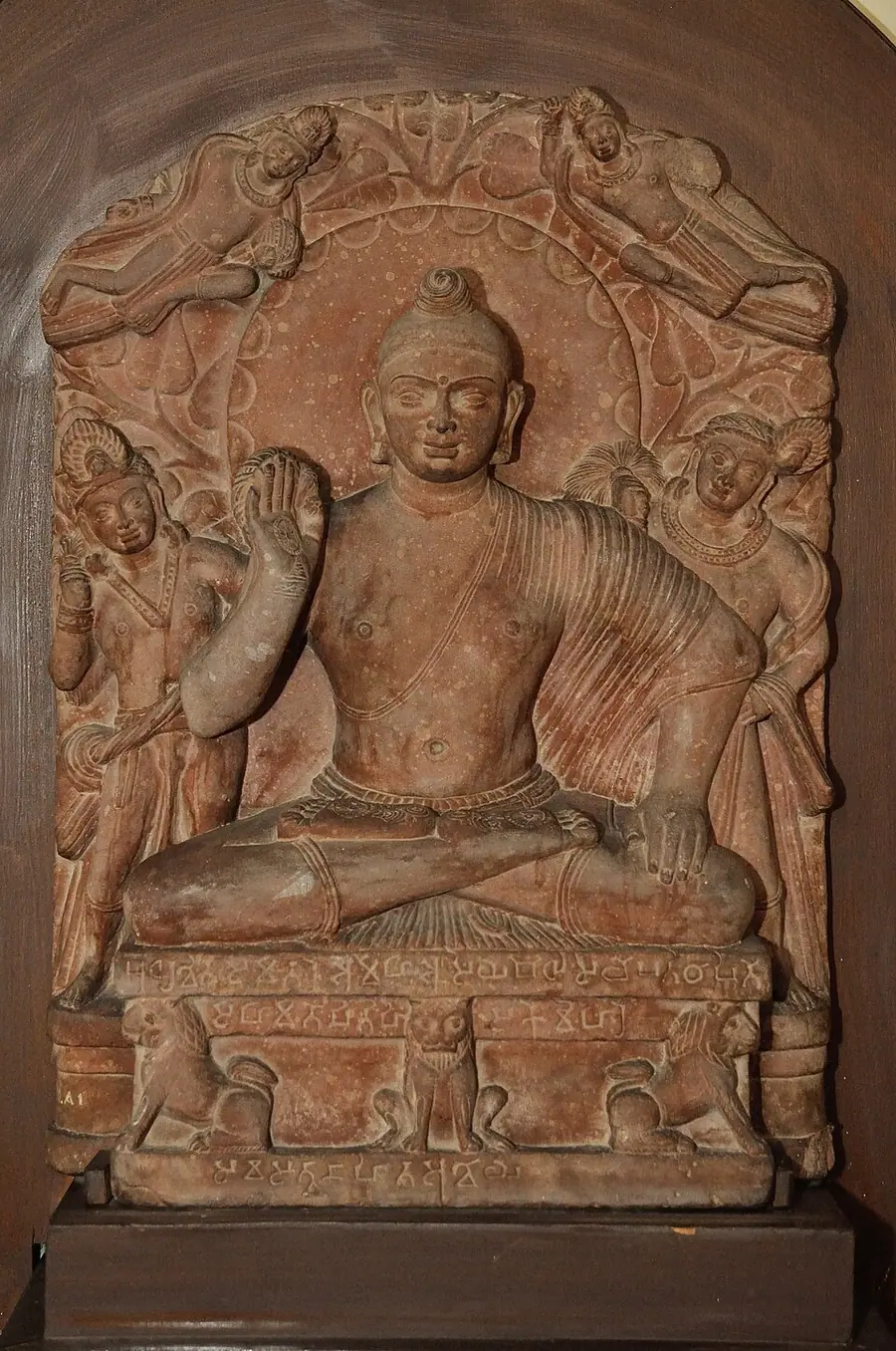 Red sandstone stele of a seated Buddha with raised right hand, two attendants with fly-whisks and two flying figures, an inscription on the base