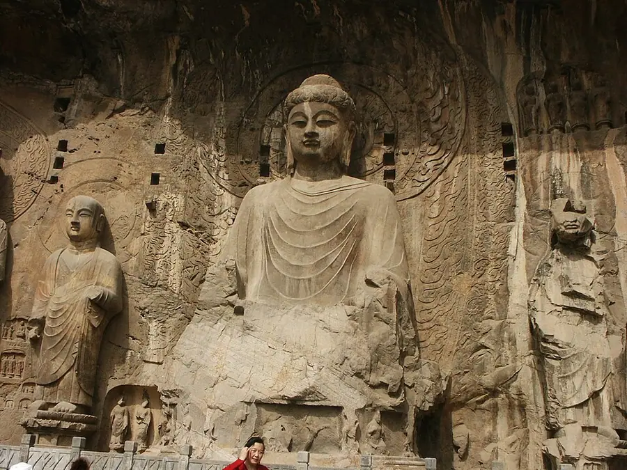 The colossal seated Vairocana Buddha carved into the cliff at Longmen's Fengxian cave, flanked by a standing monk disciple and a crowned bodhisattva