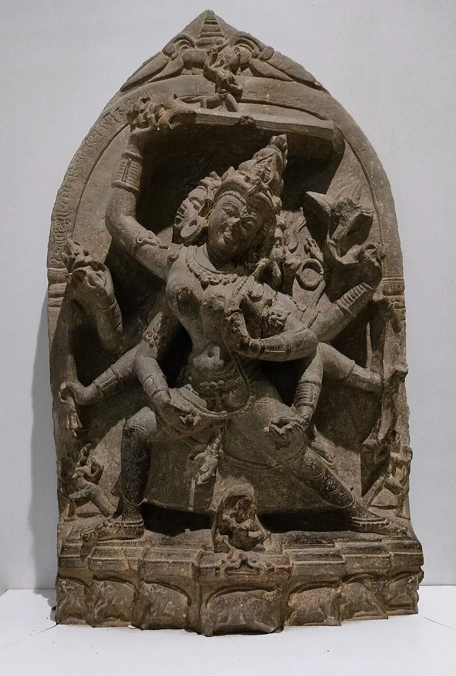 Grey stone stele of the multi-armed goddess Mārīcī in a lunging pose, with a sow's face at one side and small pigs carved along the base