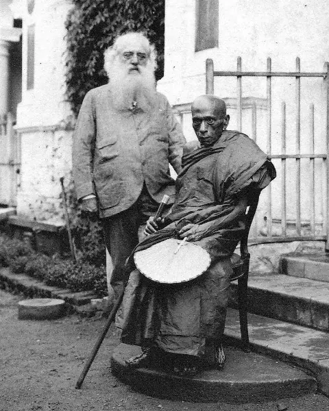 Photograph of an elderly Buddhist monk seated in robes holding a fan, with a white-bearded Western man in a suit standing beside him