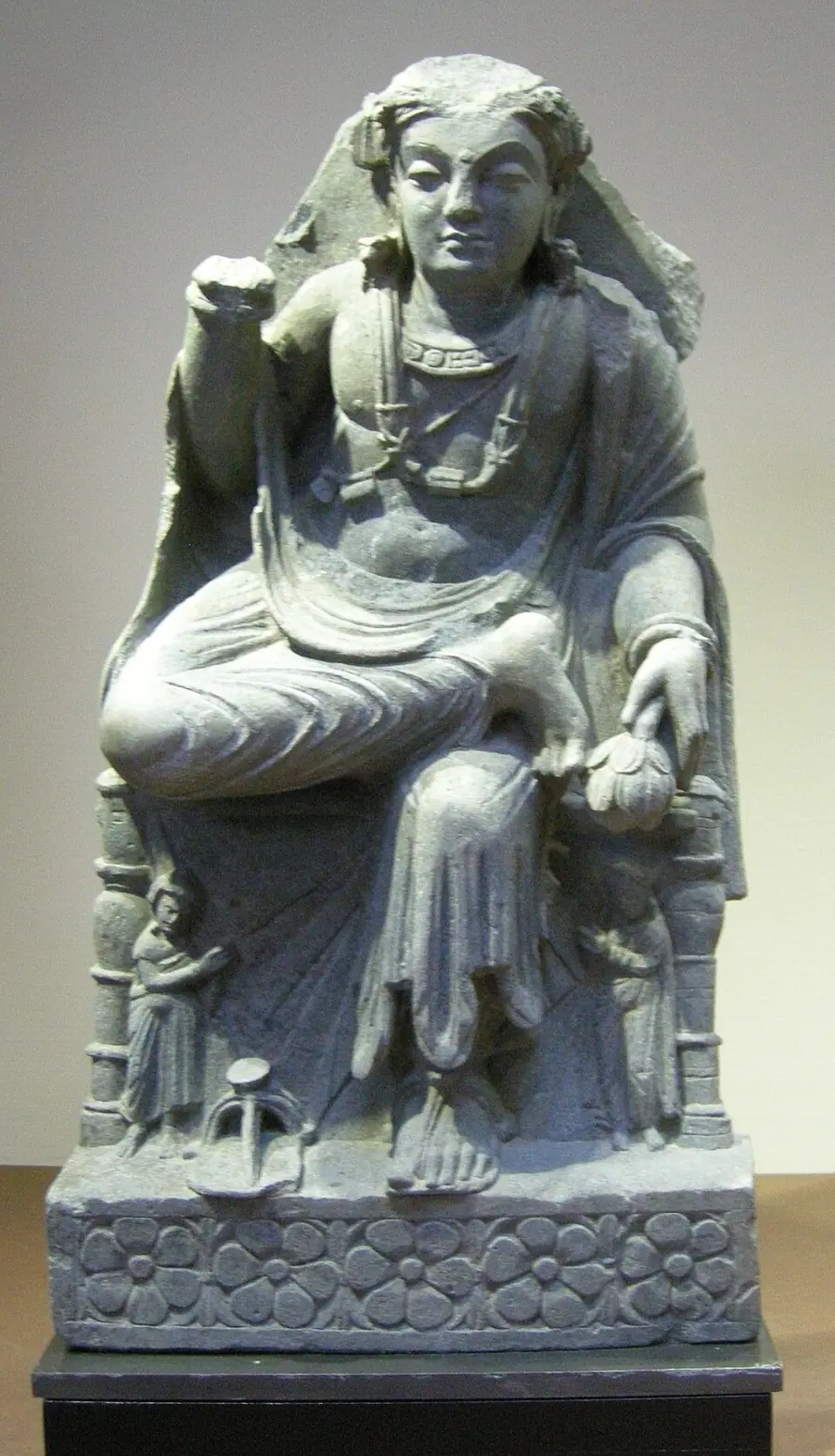 Grey schist seated bodhisattva from Gandhāra in princely dress and jewellery, holding a lotus bud, with two small attendants beside the throne