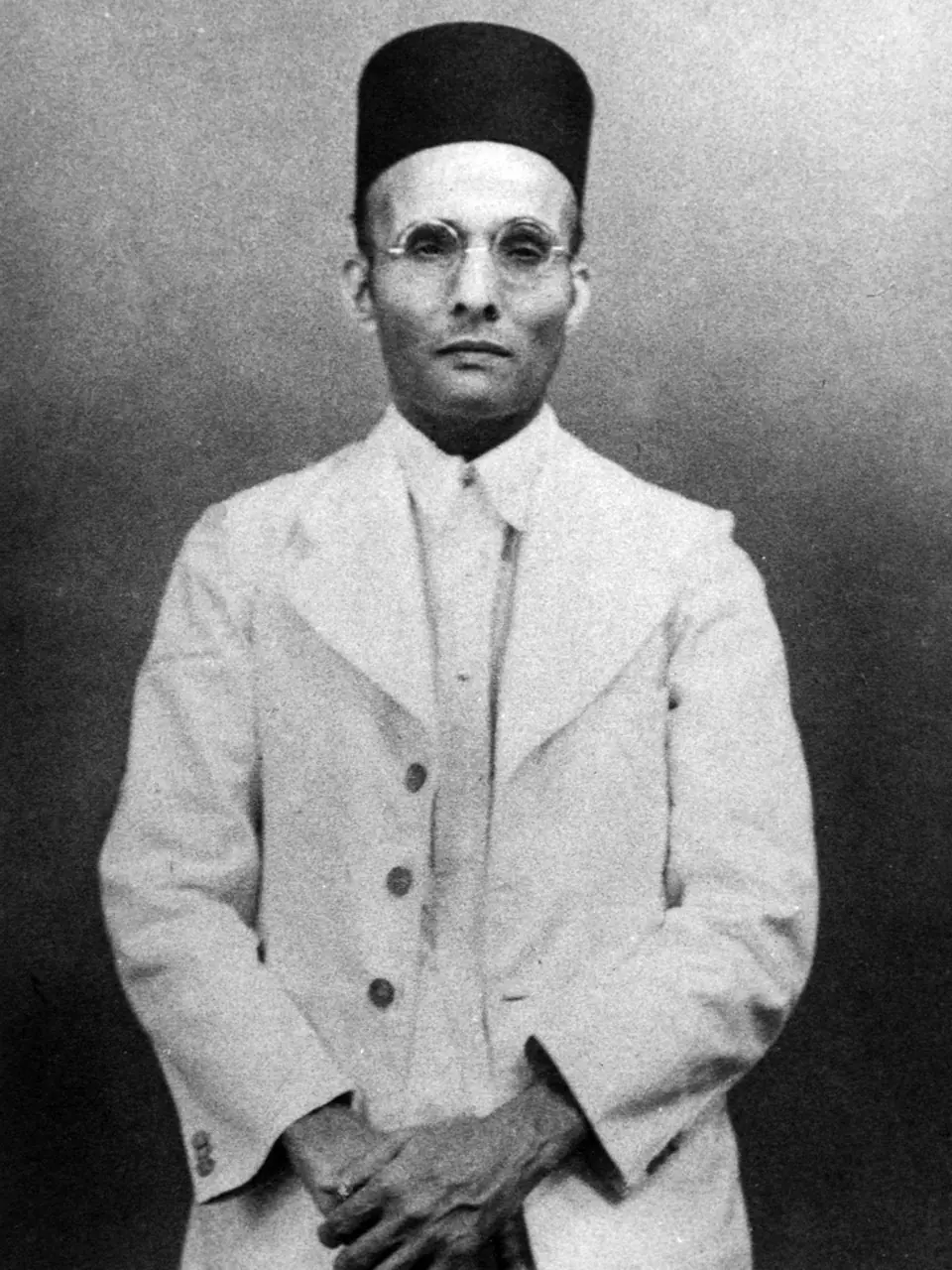 Black-and-white photograph of V. D. Savarkar in a white suit, cap and round glasses