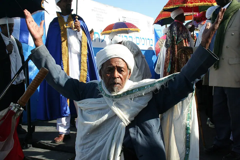 An elderly Ethiopian Jewish man in a white shawl raises his arms in prayer at the Sigd festival, with priests in embroidered robes under bright ceremonial umbrellas behind him