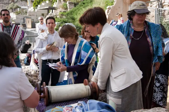 Women in prayer shawls, with a few men, gather round an open Torah scroll among the ancient stones of the Robinson's Arch area at the southern end of the Western Wall