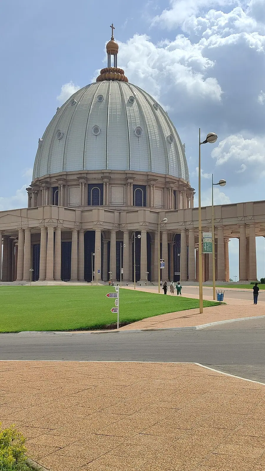 The Basilica of Our Lady of Peace at Yamoussoukro, Côte d'Ivoire, a domed church ringed by colonnades, with a few visitors walking on the lawn in front