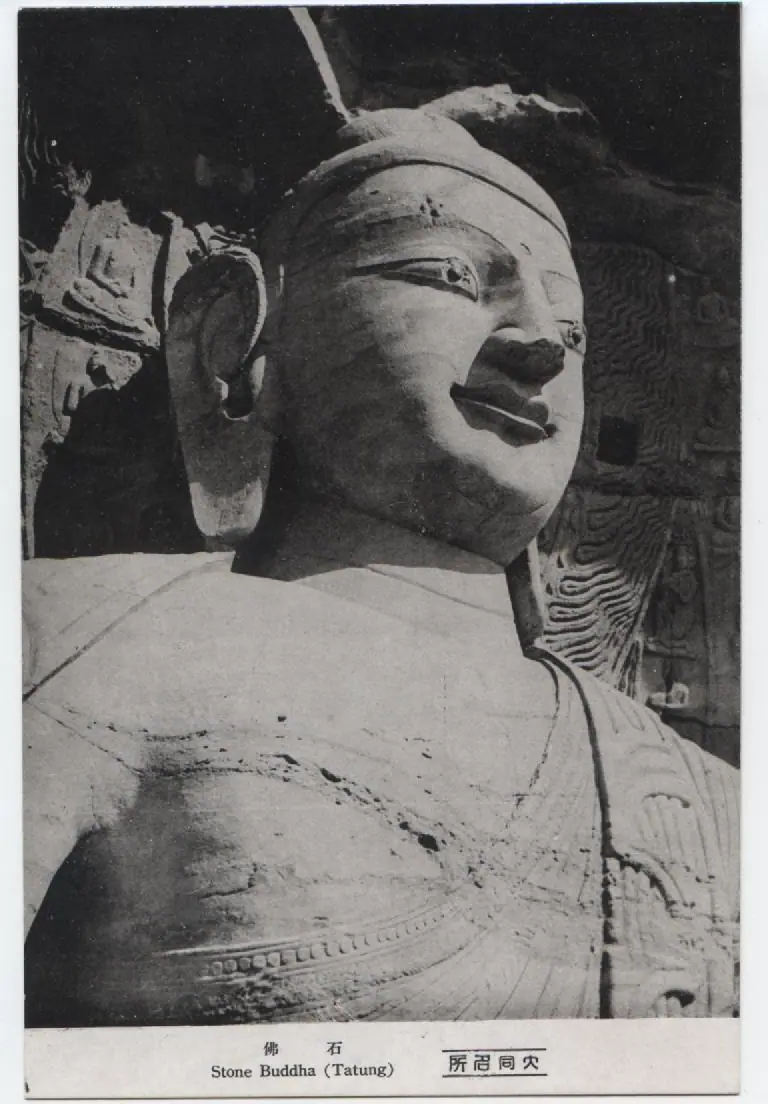 Close view of the head and shoulders of a colossal stone Buddha carved into a cave at Yungang, on an old printed postcard captioned Stone Buddha (Tatung)